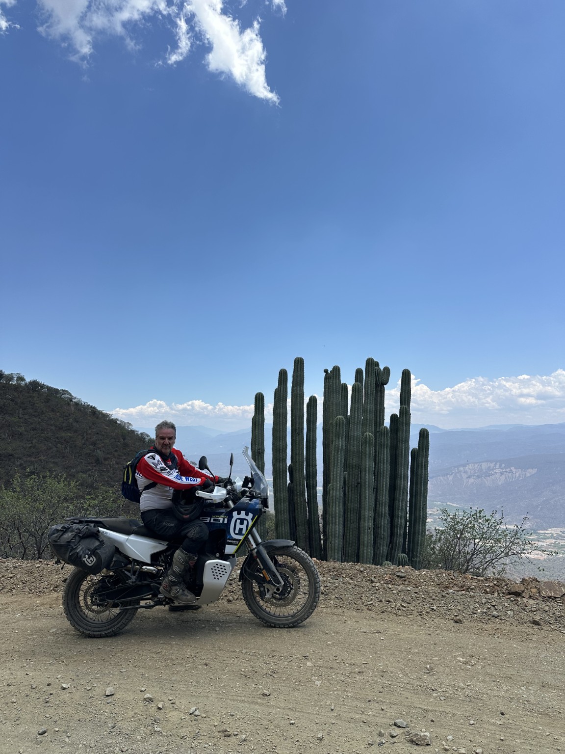 Piloto en motocicleta junto a grandes cactus con montañas al fondo